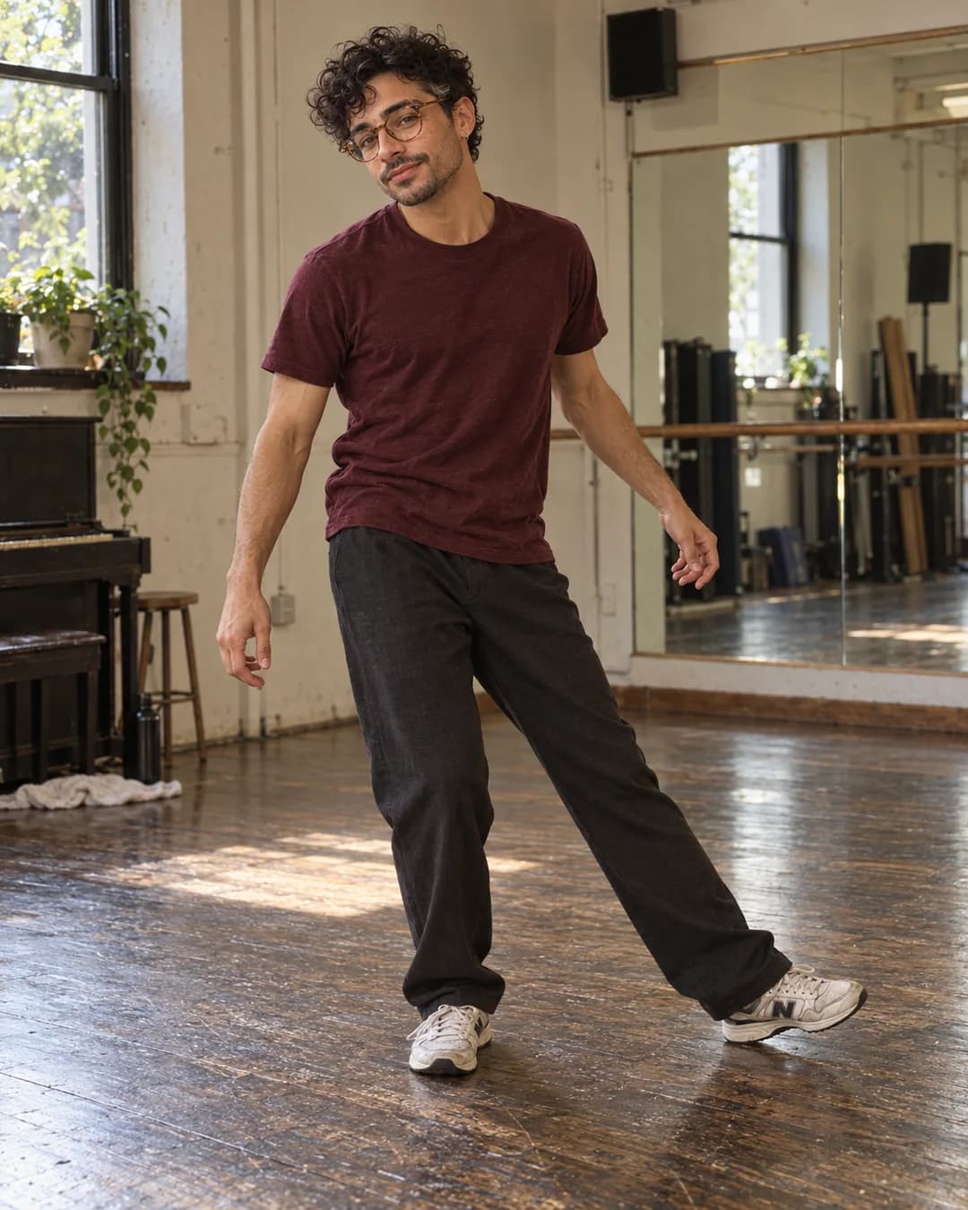 Full-body fictional Eli taking a grounded side step in a worn rehearsal studio, wearing a burgundy tee and charcoal trousers