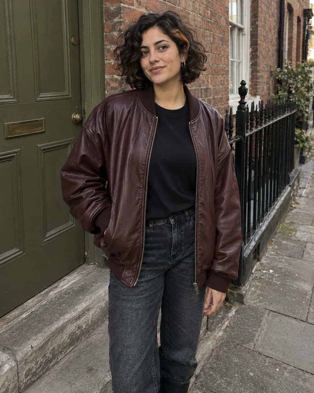 Adult virtual creator in a burgundy bomber and dark jeans beside a green door and brick wall