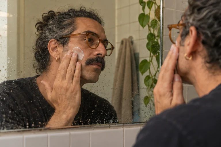 Ivo applies moisturizer to his cheek beside a bathroom mirror.