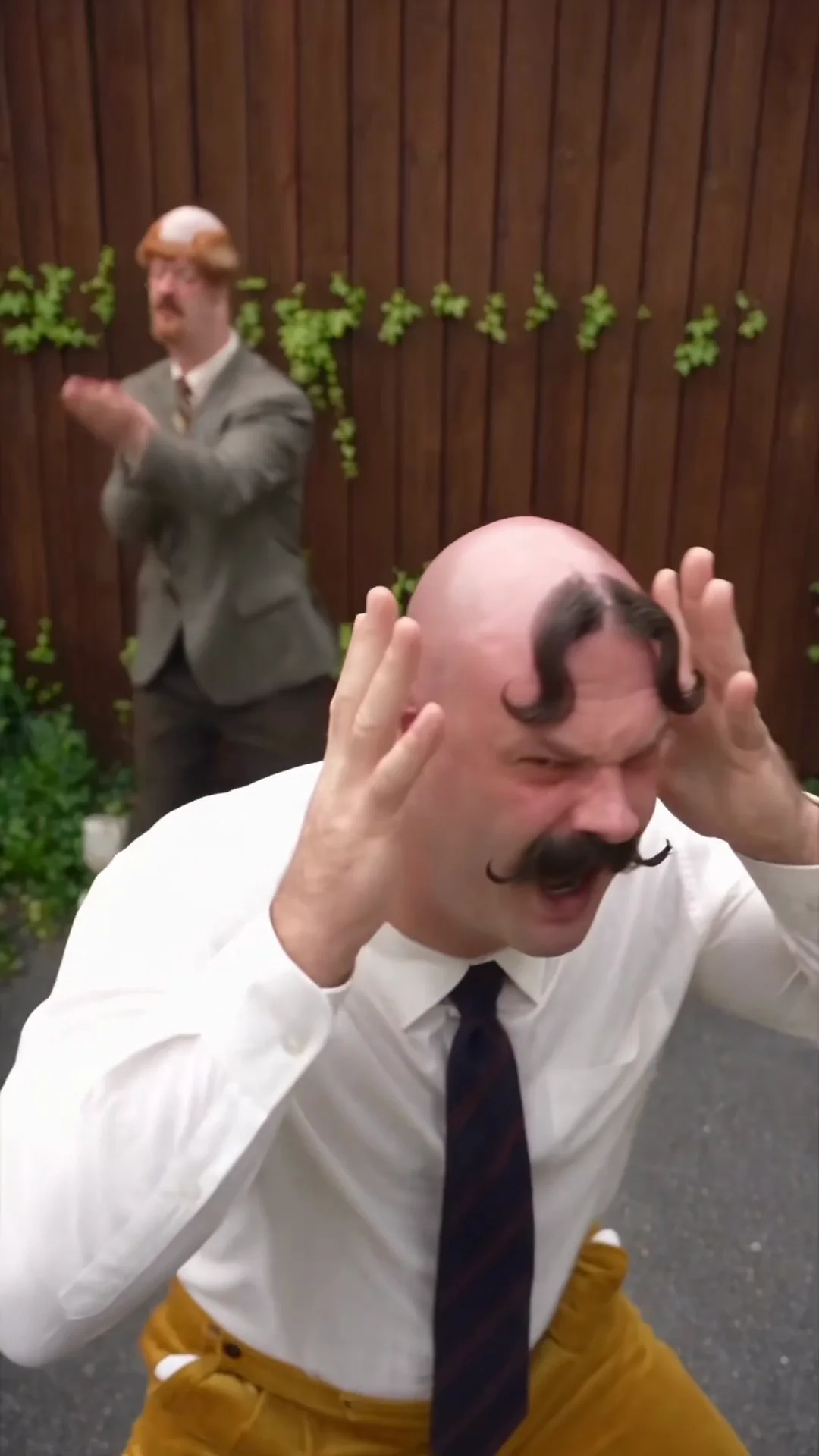 Source video at 0.5 seconds: Madman holds his head in the foreground while Harold claps near the wooden fence behind him