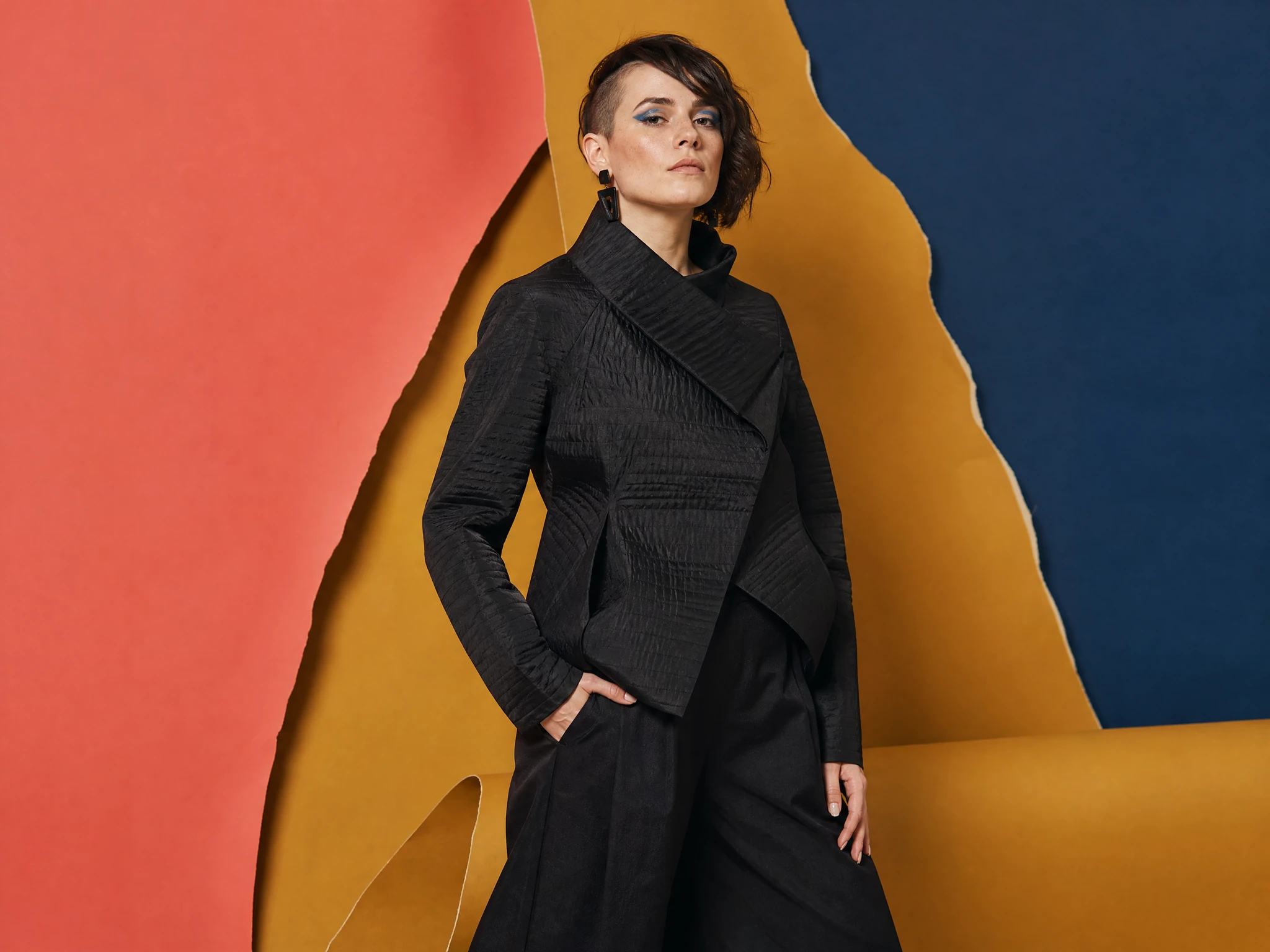 Fictional adult woman with bold graphic makeup standing against a colorful editorial backdrop.