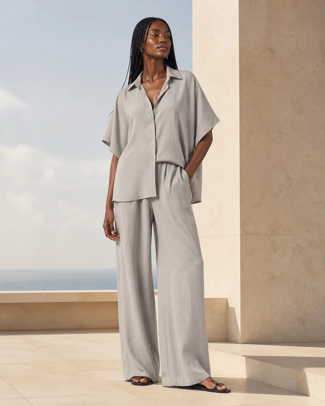 Fictional adult dark-skinned fashion model wearing an original stone-gray resort outfit on a sunlit terrace.